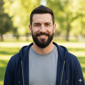 Man with a beard and mustache wearing a navy blue hoodie and gray shirt, standing in a park.