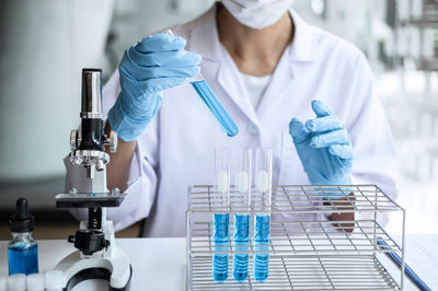 Person in a lab setting with a microscope and test tubes, wearing gloves and a lab coat.